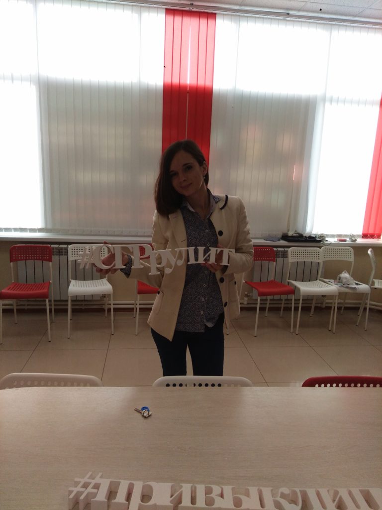 На фотографии сотрудница компании держит в руках слитную надпись, выполненную из пенопласта с хештегом